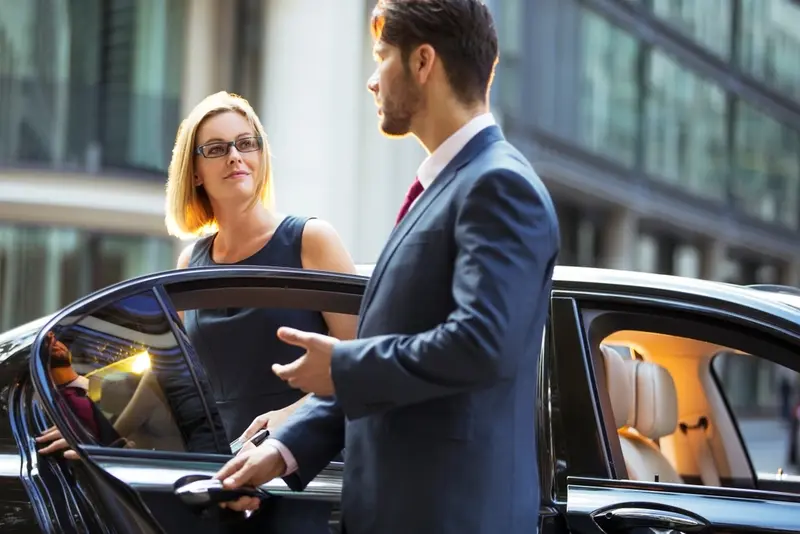 A professional chauffeur opens the rear passenger door for a stylish young businesswoman stepping out of a luxury black car—symbolizing Lavish Ride’s premium Galveston cruise shuttle service from Houston.